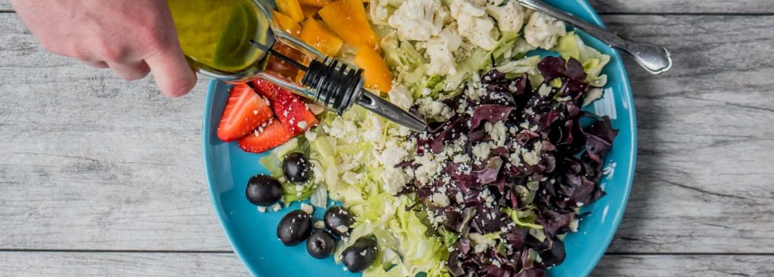 A vibrant vegetable salad with olives, feta, and strawberries being drizzled with olive oil.
